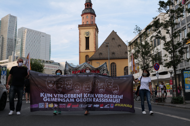 Eine Gruppe von Menschen in Masken, die eine Fahne mit Text und Bildern tragen, geht eine Straße entlang, mit einem geparkten Auto auf der linken Seite, Gebäuden, Bäumen, Schildern, Polen, einem Kirchturm und einem klaren blauen Himmel im Hintergrund.