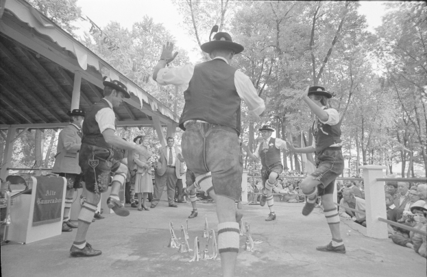 Eine Gruppe von Männern in traditioneller bayrischer Tracht tanzt auf einer Bühne vor einem gemischten Publikum, mit Bäumen und einer Hütte im Hintergrund, dargestellt in Schwarz-Weiß.