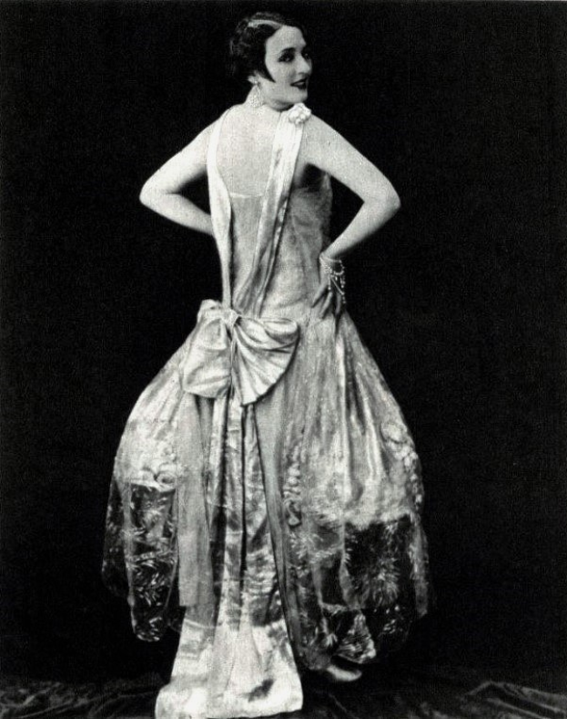 Eine Frau in einem Kleid im 1920er-Stil mit hohem Halsausschnitt, langen Ärmeln und einem voluminösen Rock posiert selbstbewusst in einem Schwarz-Weiß-Foto, ihr Haar ist elegant frisiert und das Make-up dezent.