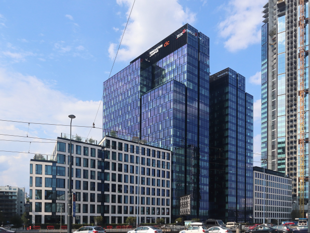 Zentrale der Deutschen Bank in Frankfurt, Deutschland, ein großes Gebäude mit vielen Fenstern, umgeben von städtischen Elementen einschließlich Straßenlaternen, Fahrzeugen, Grünflächen und einem bewölkten Himmel.