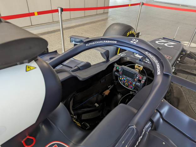 Cockpit eines Formel-E-Autos in einer Ausstellung mit einem Lenkrad im Vordergrund, einer Wand im Hintergrund und dekorativen Bändern um Pfosten.
