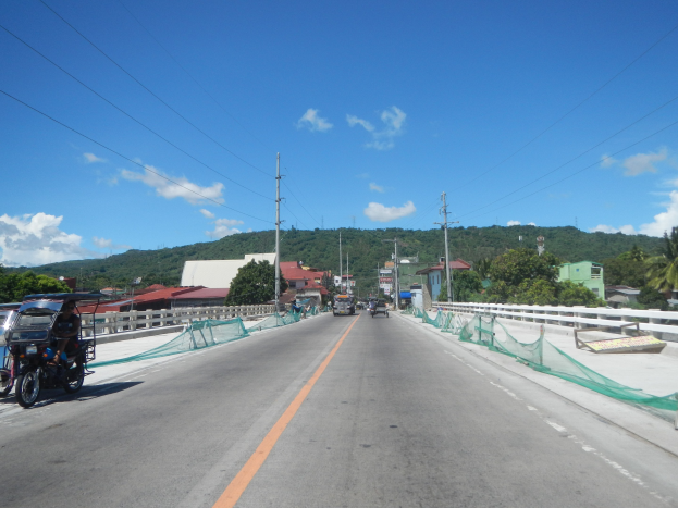 Autobahn mit fahrenden Fahrzeugen, ein geparktes Motorrad auf dem Seitenstreifen, Geländer, Strommasten mit Drähten, Bäume, entfernte Gebäude, Hügel und ein bewölkter Himmel.