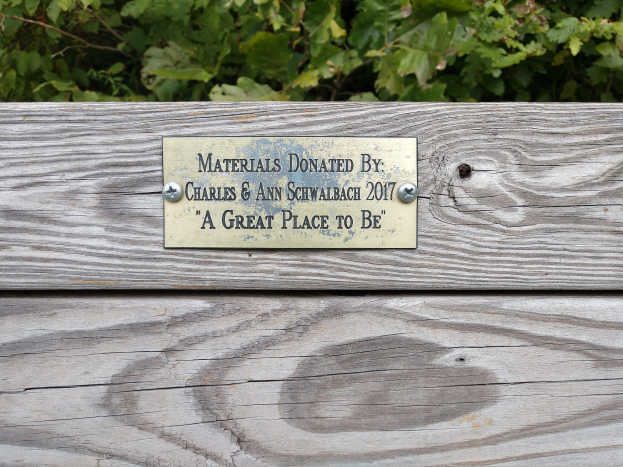 Holzbank mit einer Plakette, auf der "Materialien gespendet von Charles & Ann Schwabach 2017: Ein toller Ort zu sein" steht, vor üppigen Grünpflanzen.