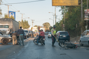 Eine Gruppe von Menschen um ein verunglücktes Motorrad auf der Straße herumstehend mit mehreren Fahrzeugen, einschließlich eines Lastwagens, und einem Hintergrund aus Bäumen, Polen, Lampen und Schildern unter einem Himmel.