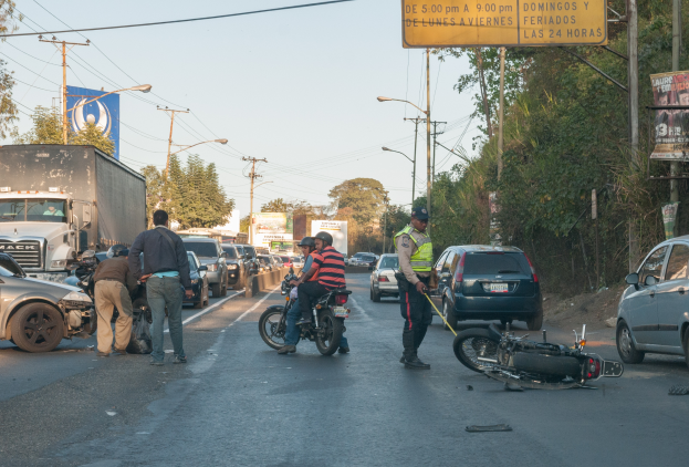 Eine Gruppe von Menschen um ein verunglücktes Motorrad auf der Straße herumstehend mit mehreren Fahrzeugen, einschließlich eines Lastwagens, und einem Hintergrund aus Bäumen, Polen, Lampen und Schildern unter einem Himmel.