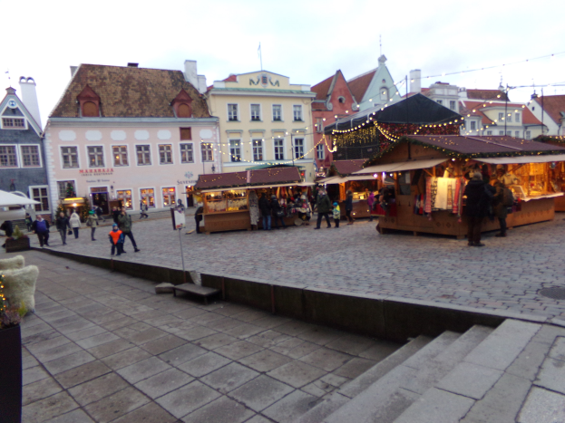 Ein geschäftiger Weihnachtsmarkt in Tallinn, Estland mit Menschen um geschmückte Stände, festlicher Beleuchtung, Gebäuden im Hintergrund, bewölktem Himmel, Treppe nach oben und Topfpflanzen.