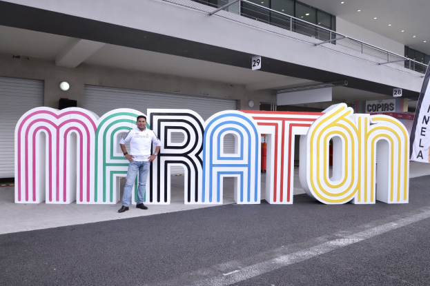 Ein Mann steht vor einem großen "Marathon"-Schild mit einem Banner rechts daneben, im Hintergrund ein Gebäude mit Lichtern, Geländern und Läden.