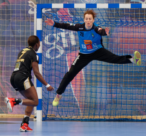 Zwei Frauen beim Handballspielen auf einem Platz mit einem Tor und einer Banner im Hintergrund.