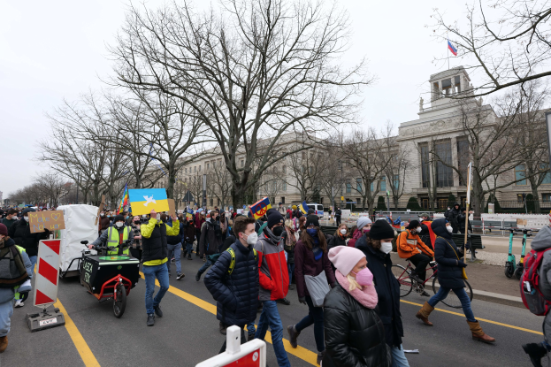 Eine große Gruppe von Menschen marschiert auf einer Straße in Washington, D.C. und hält Schilder und fährt Fahrräder, mit Bäumen, Schildern und einem klaren blauen Himmel im Hintergrund.