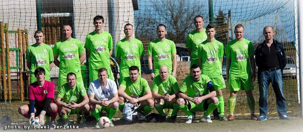 Eine Gruppe von M├Ąnnern in grünen T-Shirts steht auf einem Fussballfeld, einige sind in der Hocke, mit einem Ball vor ihnen und einem Torpfosten rechts daneben, vor einer Kulisse aus B├Ąumen, Geb├Ąuden und Fahrzeugen.