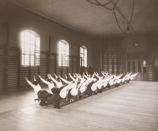 Gruppe von Menschen, die Pilates-Übungen in einem Fitnessstudio mit einer Uhr und Fenstern an der Wand im Hintergrund durchführt.