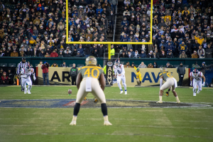 Ein Fussballspiel im Gange mit Spielern in Helmen und Sportbekleidung im Vordergrund und einer Zuschauermenge im Hintergrund, mit sichtbaren Pfählen und Texttafeln.