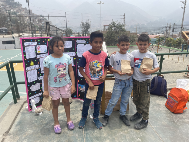 Eine Gruppe von Kindern hält Papiertüten in den Händen und steht auf einem Basketballfeld mit Tüten und Gegenständen rechts und einer Tafel mit Text links, mit einem Zaun, Pfosten und Gebäuden im Hintergrund.