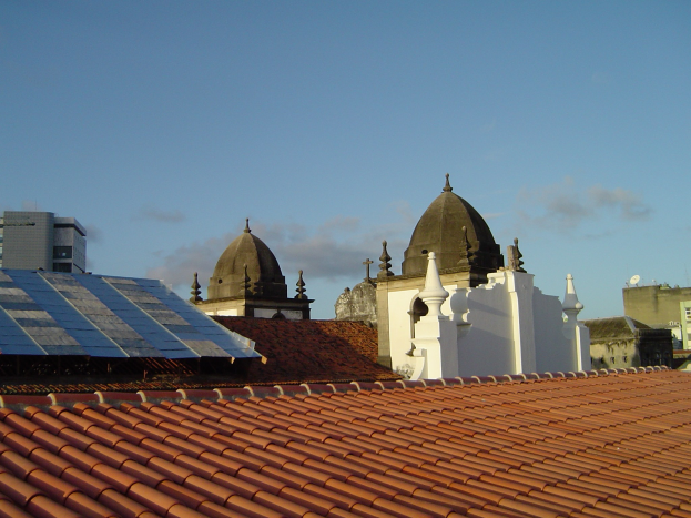 Stadtansicht mit Geb├Ąuden im Vordergrund und einem blauen Himmel im Hintergrund, mit Solarpanelen auf einem Dach, die den Einsatz von erneuerbaren Energien anzeigen.