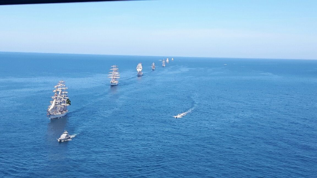 Eine große Gruppe von Segelschiffen, die auf dem Meer während eines Regattas segeln, mit einem klaren blauen Himmel und hellen Sonnenstrahlen, die auf dem Wasser reflektieren.