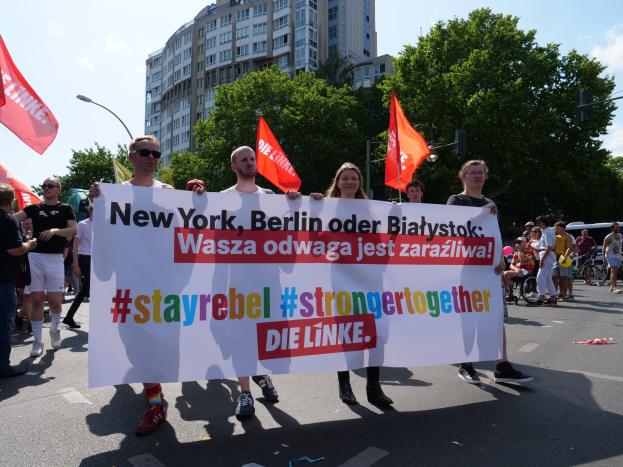 Eine Gruppe von Menschen geht eine Straße in Berlin entlang und hält ein Transparent mit der Aufschrift "Stay Rebel Stronger Together", während Bäume, Laternenpfähle und Gebäude die Straße säumen und der Himmel bewölkt ist.