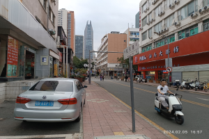 Ein Mann fährt mit einem Roller eine Straße mit hohen Gebäuden entlang, mit Menschen auf den Gehwegen, Fahrzeugen auf der Straße und Bäumen unter einem klaren blauen Himmel im Hintergrund.