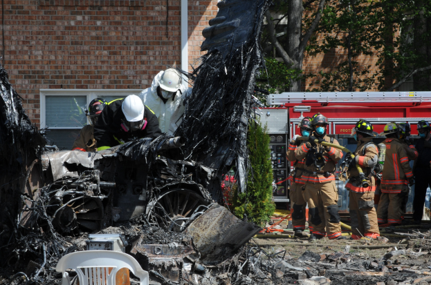 Feuerwehrleute in Schutzausrüstung löschen ein Gebäude Feuer, mit verkohlten Überresten einschließlich eines Fahrzeugs, Stuhl und Pflanzen auf dem Boden.