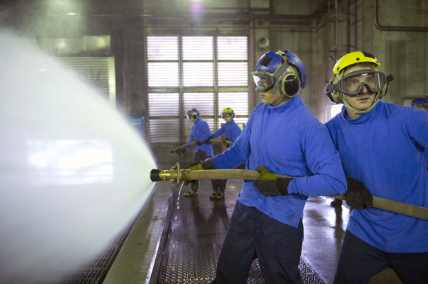 Gruppe von Männern in blauen Hemden und gelben Helmen bei der Arbeit an einer Maschine in einer Fabrik mit sichtbaren Rohren, Fenstern und Lampen.