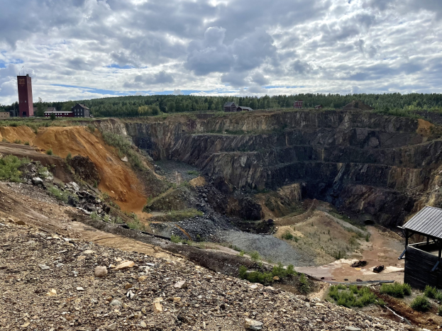 Großer Tagebau auf einem Schmutzfeld umgeben von Bäumen, Gebäuden und einem Schuppen unter einem bewölkten Himmel, mit Steinen und Pflanzen auf dem Boden.