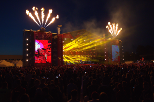 Eine große Menschenmenge auf einem Musikfestival mit einer Bühne in der Mitte, Musiker, die Instrumente spielen, unter hellen Scheinwerfern und Bannern, Zelte links daneben und ein dunkler Himmel mit Feuerwerk im Hintergrund.