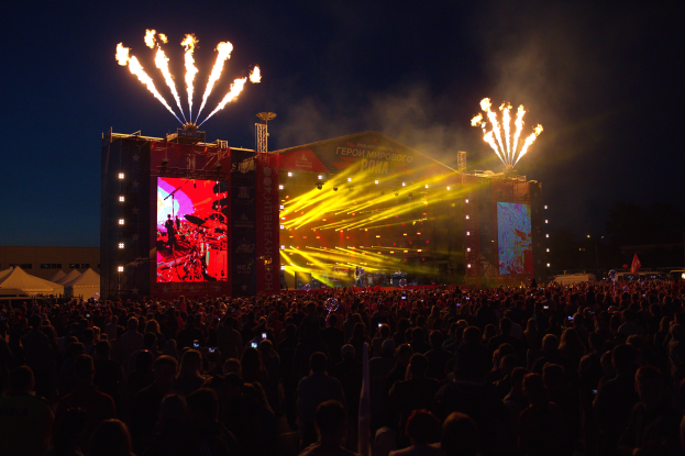 Eine große Menschenmenge auf einem Musikfestival mit einer Bühne in der Mitte, Musiker, die Instrumente spielen, unter hellen Scheinwerfern und Bannern, Zelte links daneben und ein dunkler Himmel mit Feuerwerk im Hintergrund.