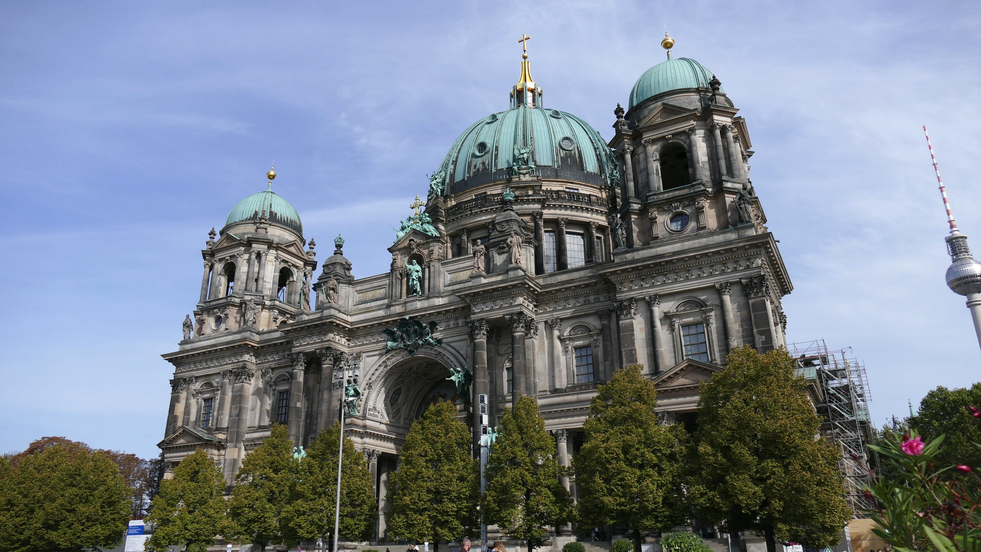 Großes, prunkvolles Berliner Dom-Gebäude in Berlin, Deutschland, mit architektonischen Details wie Fenstern, Säulen, Bögen und Statuen, vor einem bewölkten Himmel mit Menschen, Bäumen und einem Turm im Vordergrund.