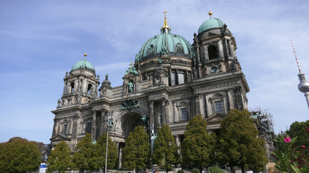 Großes, prunkvolles Berliner Dom-Gebäude in Berlin, Deutschland, mit architektonischen Details wie Fenstern, Säulen, Bögen und Statuen, vor einem bewölkten Himmel mit Menschen, Bäumen und einem Turm im Vordergrund.