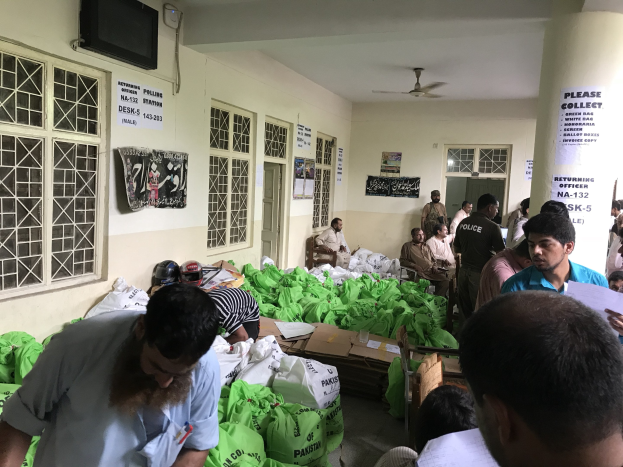 Menschen in einem Raum mit Lebensmittelbeuteln, einige sitzen auf Stühlen und andere stehen, einige halten Papiere, mit Fenstern, Plakaten, einem Fernseher, Türen, einer Säule und Deckenventilatoren.