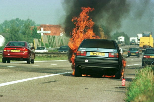 Ein Auto, das von Flammen umgeben ist, am Straßenrand mit anderen Fahrzeugen in der Nähe, umgeben von Bäumen, Gebäuden und einem klaren blauen Himmel, mit Gras und einem Feuerlöscher auf der rechten Seite.
