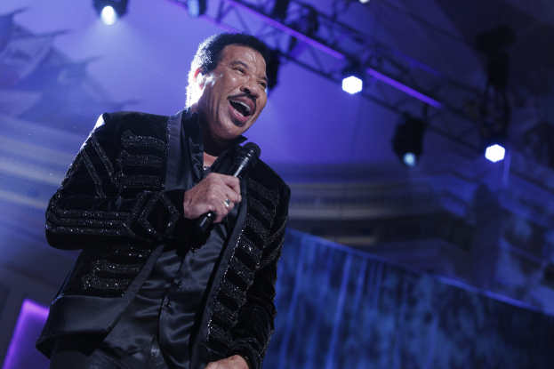Lionel Richie performs auf den Grammys, trägt ein schwarzes Kleid und hält ein Mikrofon auf einer hell erleuchteten Bühne.