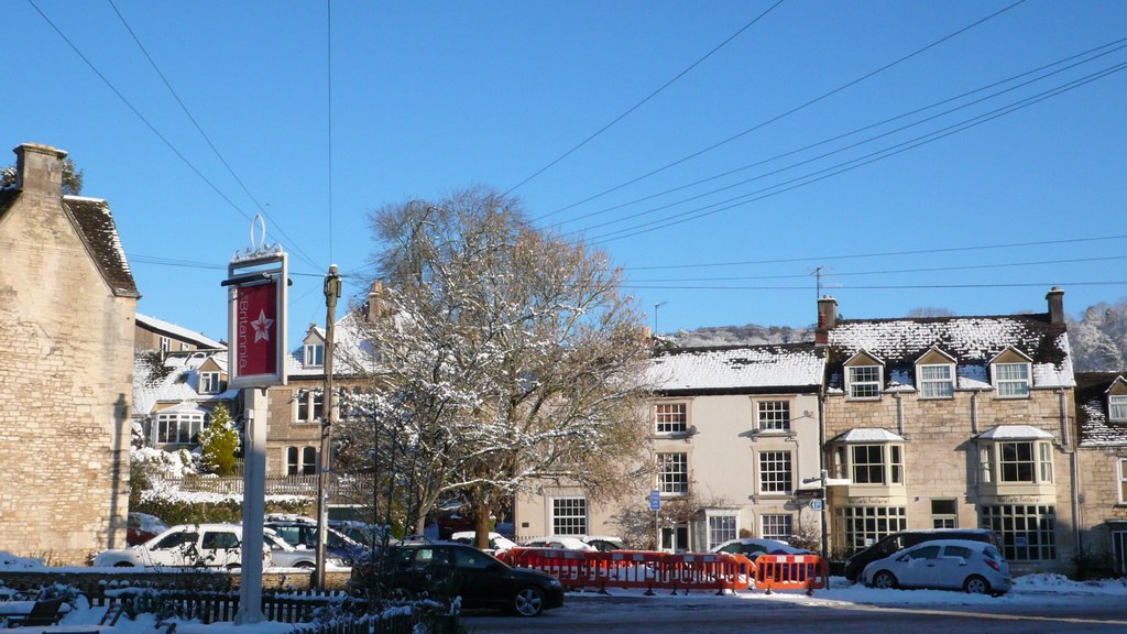 Schneebedeckte Fahrzeuge auf einer Straße mit Häusern, Bäumen, Strommasten und schneebedeckten Drähten im Hintergrund.