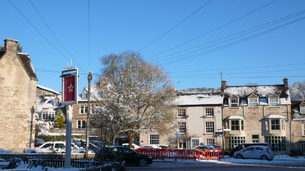 Schneebedeckte Fahrzeuge auf einer Straße mit Häusern, Bäumen, Strommasten und schneebedeckten Drähten im Hintergrund.