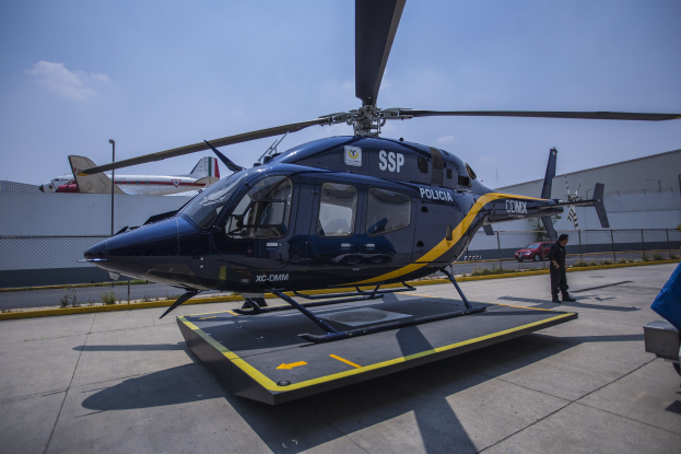 Polizeihubschrauber auf dem Rollfeld mit einer Person davor, Flugzeuge und ein Auto im Hintergrund, umgeben von einem Zaun, Pflanzen und einem klaren blauen Himmel.