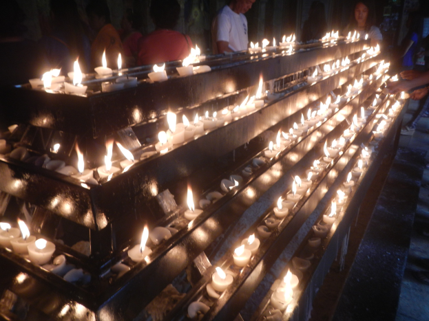 Eine Gruppe von Menschen steht um eine lange Reihe von brennenden Kerzen in einer Kirche, erhellt von ihrem flackernden Licht gegen eine Wand.