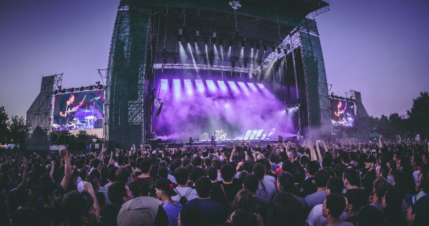 Eine große Menge bei einem Musikfestival mit einer Bühne, auf der Musiker spielen, beleuchtet von Lichtern und umgeben von Bildschirmen, vor einer Kulisse aus Gebäude, Bäumen und einem klaren blauen Himmel.
