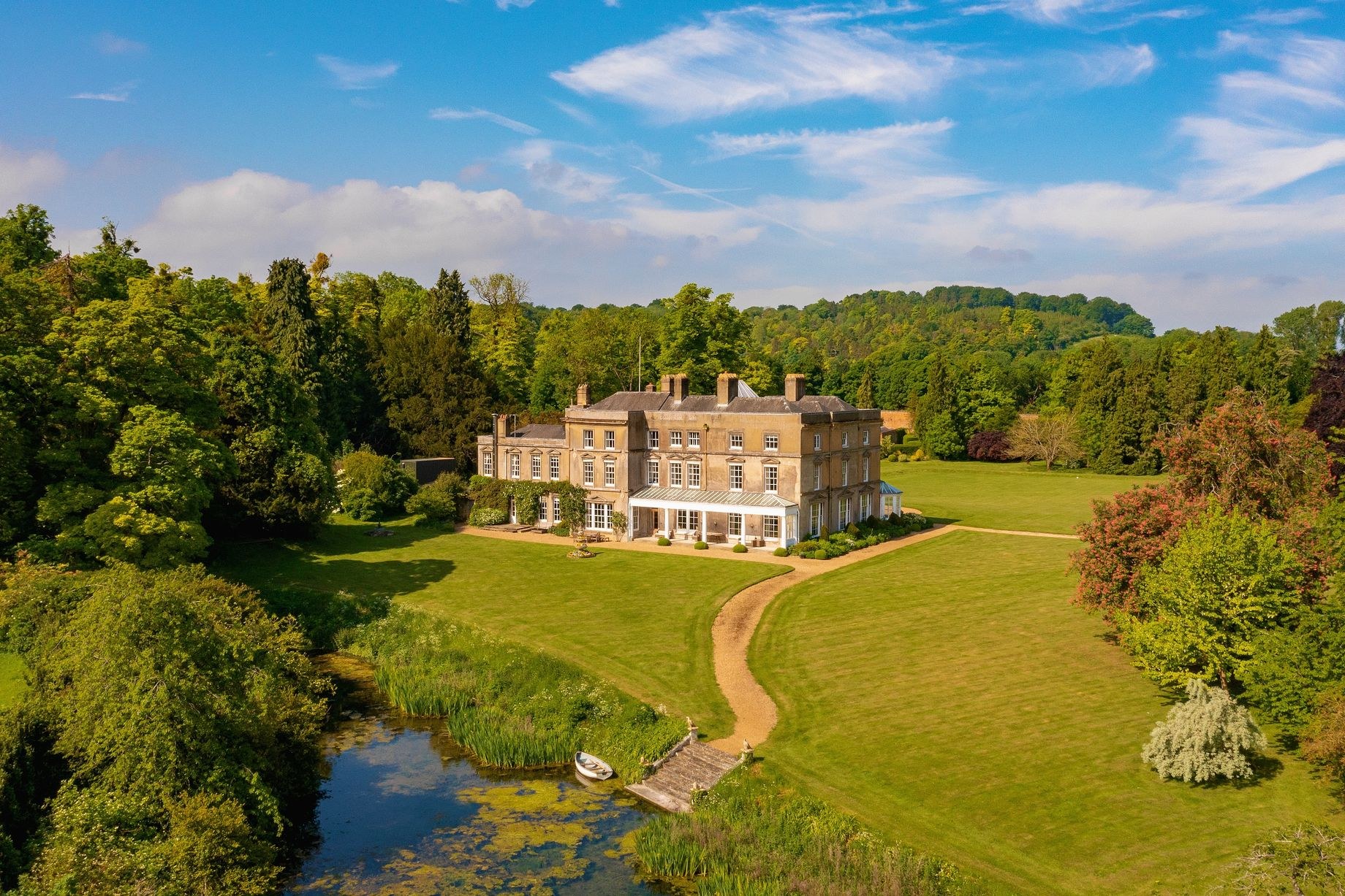 Luftaufnahme eines großen, teuren Anwesens in einer grünen Wiese mit einem Weg, umgeben von Bäumen und Pflanzen, einem Gewässer im Vordergrund und bewölktem Himmel.
