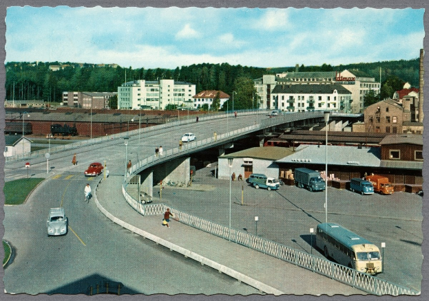 Altes Schwarz-Weiß-Foto einer Stadtstraße mit fahrenden Autos und Bussen, Fußgängern auf einer Brücke, Laternen, mehrstöckigen Gebäuden mit Fenstern, Bäumen und einem bewölkten Himmel.