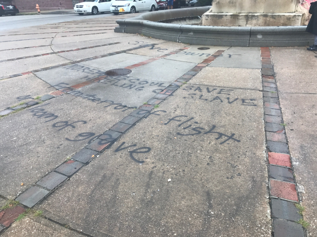 Geweg mit 'Black Lives Matter'-Graffiti in großen Buchstaben, umgeben von einem Gebäude, Fahrzeugen, Menschen, Bäumen, Pfählen und einer Wand.