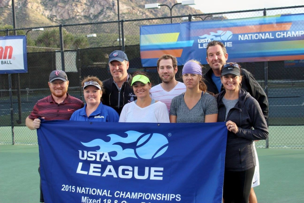 Gruppe von Menschen, die auf einem Tennisplatz mit einem Zaun, Bannern, Laternenmasten, Bäumen, Bergen und einem klaren blauen Himmel im Hintergrund eine "2015 USTA National Championships"-Tafel halten und lächeln