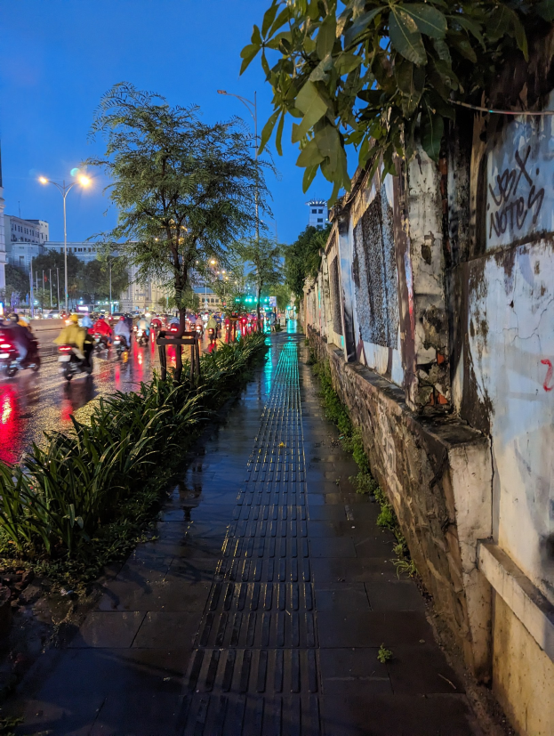 Stadtstraße bei Nacht mit Graffiti an der Seite, beleuchtet von Straßenlaternen, fahrende Kraftfahrzeuge, Bäume und Pflanzen an den Seiten, Gebäude und ein sternenübersäter Himmel im Hintergrund.