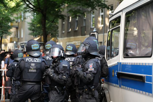 Eine Gruppe von Polizeibeamten vor einem Bus auf einer Straße mit einer Menge hinter Absperrungen, Bäumen und Gebäuden im Hintergrund.