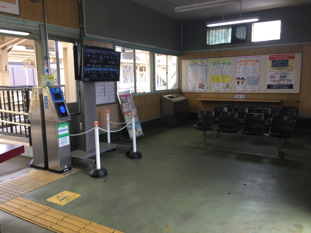 Ein belebter Bahnhofseingang mit Reihen von Stühlen, einem Service-Schalter, Ticketautomaten, Kettengliedern, Informationsanzeigen, Wandplakaten, Deckenbeleuchtung und Geländern.