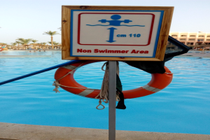 Ein Schwimmbad mit einem Schild "Nichtschwimmerbereich" und einem Rettungsring an einem Pfahl, vor Bäumen, Gebäuden und einem klaren blauen Himmel.