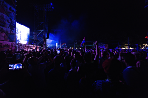 Eine Menschenmenge auf einem Konzert mit erhobenen Händen, einige halten Mobiltelefone und Fahnen, vor einem Bildschirm mit Bildern und Text, Metallrahmen und Lichtern vor einem dunklen Hintergrund.