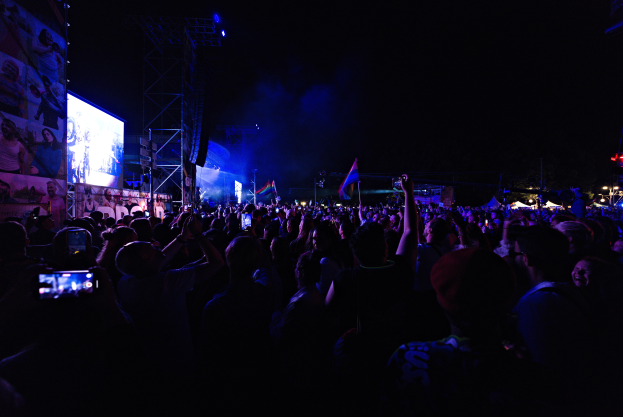 Eine Menschenmenge auf einem Konzert mit erhobenen Händen, einige halten Mobiltelefone und Fahnen, vor einem Bildschirm mit Bildern und Text, Metallrahmen und Lichtern vor einem dunklen Hintergrund.