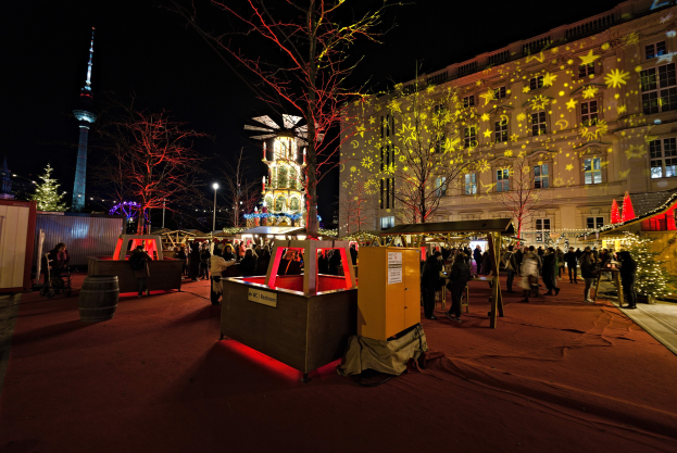 Ein geschäftiger Weihnachtsmarkt in Berlin, Deutschland mit Menschen um geschmückte Stände, festliche Lichter, Bäume, Gebäude, Laternenmasten und einen Turm unter einem dunklen Himmel.