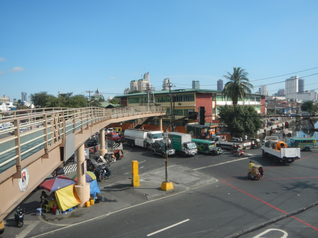 Vielbefahrene Straße mit starkem Verkehr, Fußgängern, Zelten und Gebäuden in der Nähe einer Brücke, mit Wasser und bewölktem Himmel im Hintergrund.