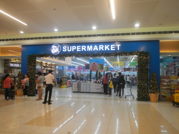 Menschen vor einem Supermarkt mit Einkaufstaschen, einigen mit einem Einkaufswagen, Topfpflanze, Regalen, Schildern und Deckenleuchten im Hintergrund.