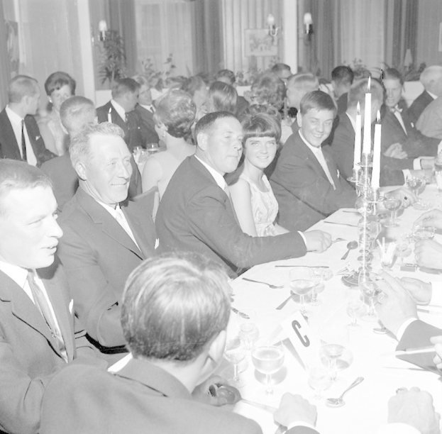 Eine Gruppe von Menschen sitzt um einen langen Tisch und lächelt, während sie die Gesellschaft des anderen genießt, mit Gläsern, Besteck, Kerzen und anderen Gegenständen auf dem Tisch, vor einem Hintergrund aus Vorhängen und Wandlampen.